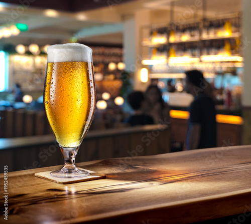 Glass of chilled beer on wooden bar tabletop and blurred brightly lit bar interior in the background.