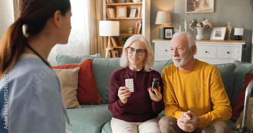 A home health nurse talks to an elderly couple in their living room. The nurse discusses medications while the couple listens, sitting on their couch.