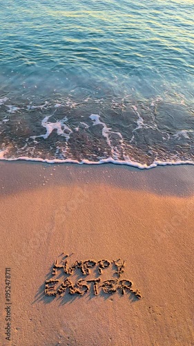 Vertical slow motion of Happy Easter written on sandy beach with gentle waves and warm sunlight