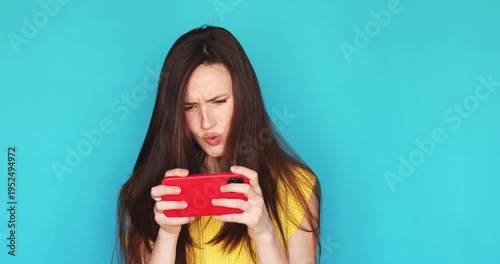 Frustrated young woman playing mobile game on smartphone in horizontal mode feeling angry losing or facing internet lag looking up with disappointment isolated on blue background mobile gaming concept