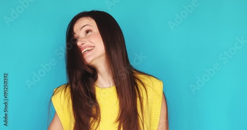 Cheerful young woman with long dark hair wearing yellow knitted vest looking at camera and laughing sincerely isolated on blue background, expressive facial emotions, happy female portrait