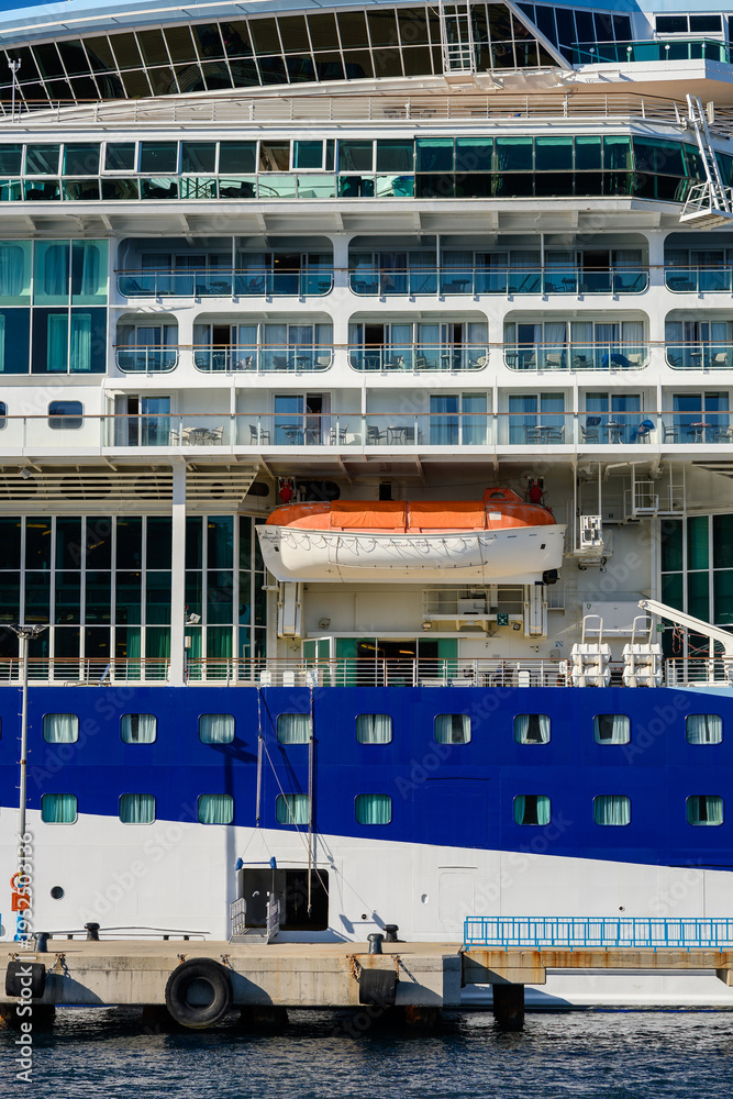 Fototapeta premium Close-up of cruise ship's hull, showing lifeboat, rows of balconies, mooring ropes, and dockside vehicler, in bright sunlight. Sea travel concept.