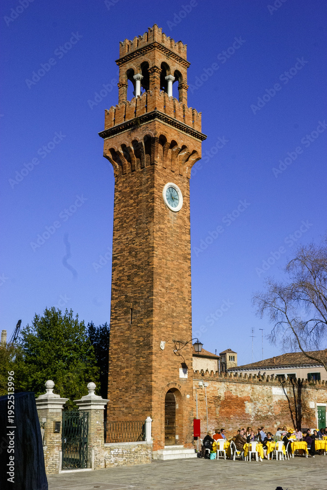 Fototapeta premium The Clock Tower in Murano, Venice, Italy, with people dining outdoors under a clear blue sky.