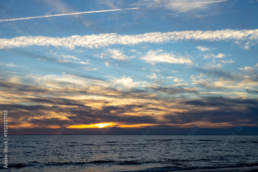 Obraz premium Scenic sunset over Mediterranean sea with distant Taurus mountains and contrails in cloudy sky. Beautiful coastal landscape with airplane trails during winter evening in Antalya region. Turkey.