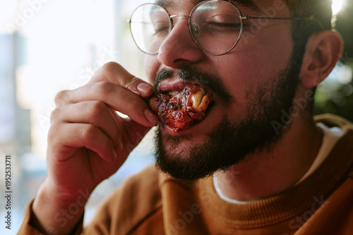 Принти на полотні Young adult biracial man eating grilled chicken wing, holding food close to mout