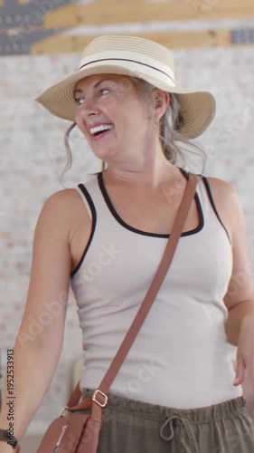 Vertical video: Entering woman smiling after greeting, hand fixing strap, moving into room with hat