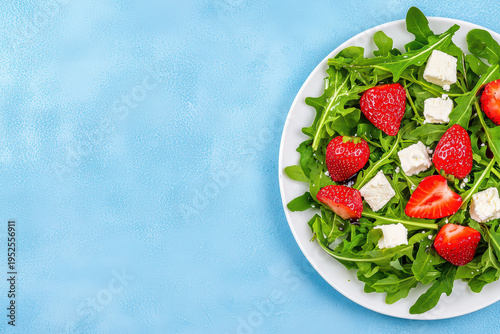 Fresh salad with arugula, strawberries, and white cheese cubes on white plate, set against textured light blue background, creating vibrant and healthy meal presentation