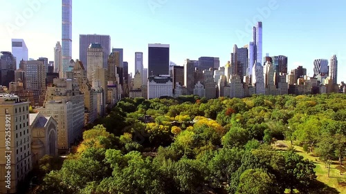 Sweeping aerial drone footage captures the lush greenery and vibrant autumn foliage of Central Park in New York City, foregrounding the iconic Manhattan skyline with ultra-tall skyscrapers.