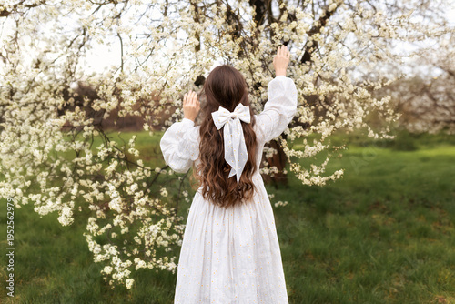 Wallpaper Mural Back view of a young woman with long hair and a white bow, standing in a blooming orchard and reaching toward delicate spring blossoms. Concept of spring, beauty, femininity, and harmony with nature. Torontodigital.ca