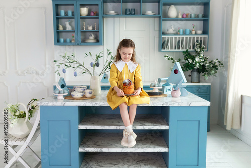 Wallpaper Mural Young girl sit on kitchen with cake, spring decor, flowers, easter eggs and bunny. Child Girl in yellow dress holding easter cake on plate in hands. Little Girl preparing festive pastries at home. Torontodigital.ca
