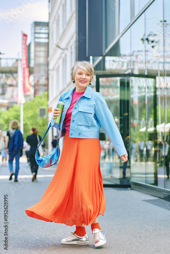 Stylish woman in her fifties with grey hair walking in modern city street, confident mature female in bright outfit holding coffee, active urban lifestyle and fashion portrait outdoors