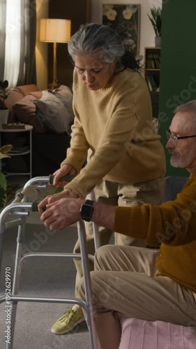 Vertical view of senior woman bringing walking frame for injured man and helping him stand up from couch in home setting