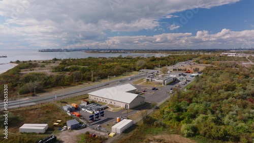 Aerial drone view of an industrial facility with warehouses, trucks, and equipment near a coastal landscape with water and greenery under a cloudy sky.