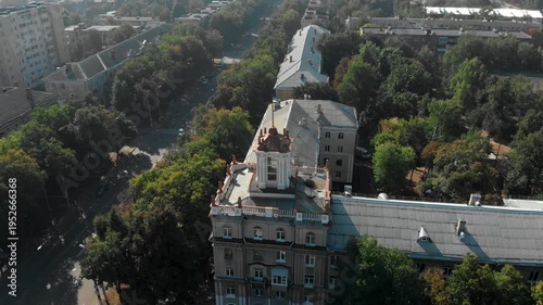 Aerial drone view of Sumy city center in Ukraine featuring historic architecture with a classical building topped by a Ukrainian flag, surrounded by green trees and urban streets.