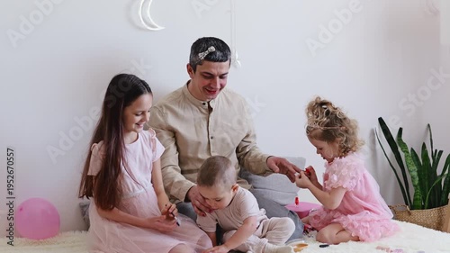 A man sits on a rug while children play with toys. One child paints his nails, and another gets her hair styled. The room has balloons and a plant, creating a lively setting
