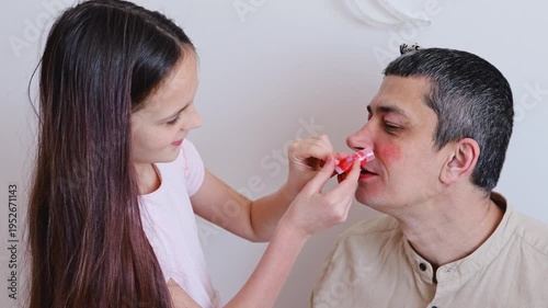 A young girl smiles as she gently touches her father's face. They share a moment in a bright room with a light background. The atmosphere shows joy and connection