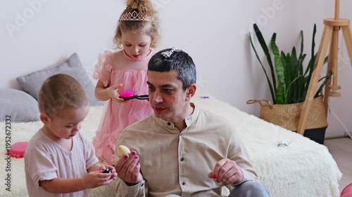 Two young children are playing with toys while a man sits on a soft surface. The room has plants and light colors, creating a playful mood