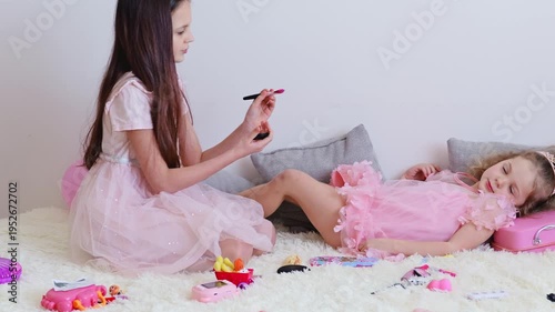 A child sits on the floor while another applies makeup. They are surrounded by toys, accessories, and colorful decorations during playtime at home