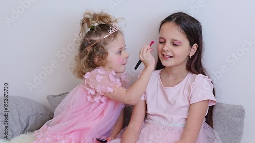 Two young girls enjoy a fun moment together as one applies makeup on the other while sitting on a sofa in a well-lit room during the afternoon