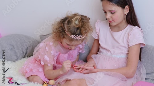 Girls sit on a soft surface in a home setting. One girl paints the nails of the other while surrounded by toys and makeup items. They appear focused and engaged in the activity