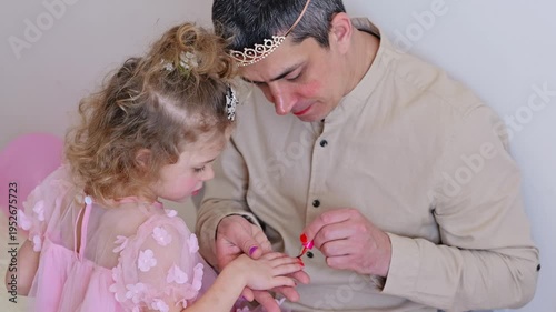 A father and his young daughter sit together, having fun as he paints her nails with red polish. They both smile and share a joyful moment in their home