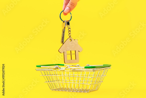 Hand holds the keys with wooden house keychain, putting them in shopping cart on bright yellow background. Purchase of apartments, mortgage, sale, purchase, and rental of real estate business.