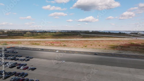 Aerial drone view of an industrial facility with warehouses, trucks, and equipment near a coastal landscape with water and greenery under a cloudy sky.