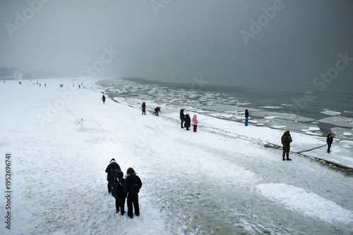Winter landscape of a frozen Baltic Sea in Sopot, Poland, with ice floes on the water and people walking on the snowy beach under a foggy sky