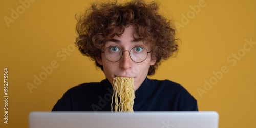 Young caucasian male ramen noodles laptop mouthful glasses chopsticks curly hair yellow background candid expression