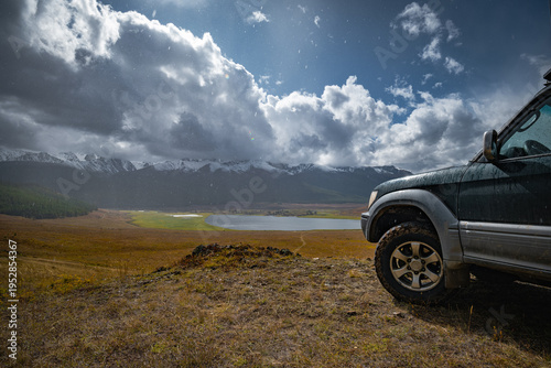 Off-road 4x4 SUV wheel and mud tire on edge of mountain plateau overlooking scenic highland lake and snow peaks during rain and sun for overland adventure journey in remote wilderness.
