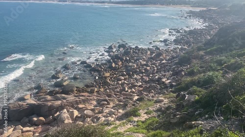 Cinematic wide shot of a rocky coastline in Vietnam with turquoise ocean waves, traditional fishing boats, and a distant wind farm under blue sky, representing coastal beauty and renewable energy.