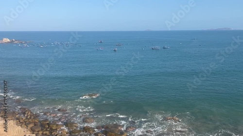 Cinematic wide shot of a rocky coastline in Vietnam with turquoise ocean waves, traditional fishing boats, and a distant wind farm under blue sky, representing coastal beauty and renewable energy.