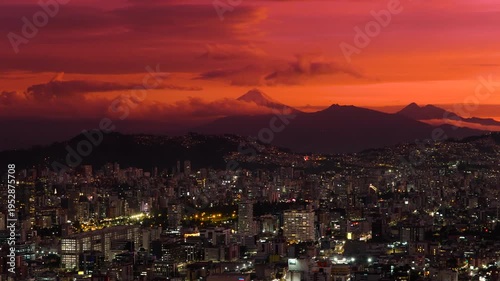 Aerial drone view of Quito, Ecuador at sunset with a dramatic orange sky, illuminated urban skyline, and Cotopaxi volcano rising in the distance above the Andes.