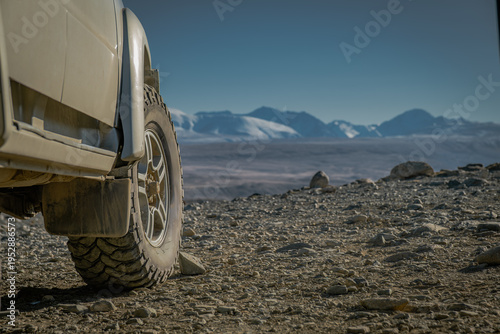 Close up of off-road 4x4 SUV wheel and mud tire on rocky edge of mountain plateau overlooking scenic snow peaks and highland valley during overland adventure journey in wilderness.
