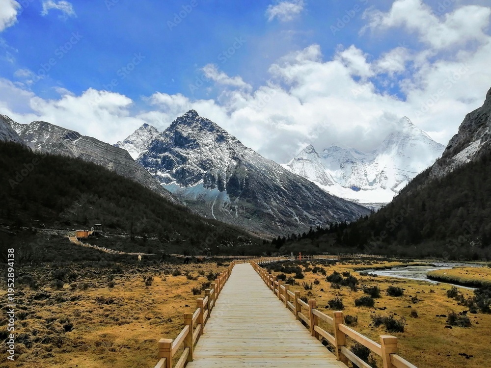Naklejka premium Wooden Boardwalk Leading to Snow-Capped Mountains in Yading Nature Reserve, Sichuan, China. Majestic alpine landscape with clear blue sky and white clouds.