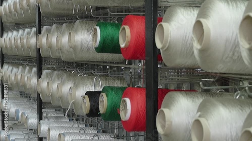 Rows of white and colored industrial yarn bobbins on a metal rack in a textile manufacturing factory.