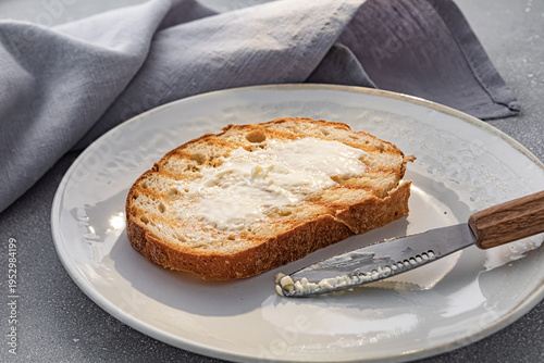 Freshly toasted artisan bread with melting creamy butter on a white ceramic plate. Close-up of bread texture with grill marks and a wooden handle butter knife.