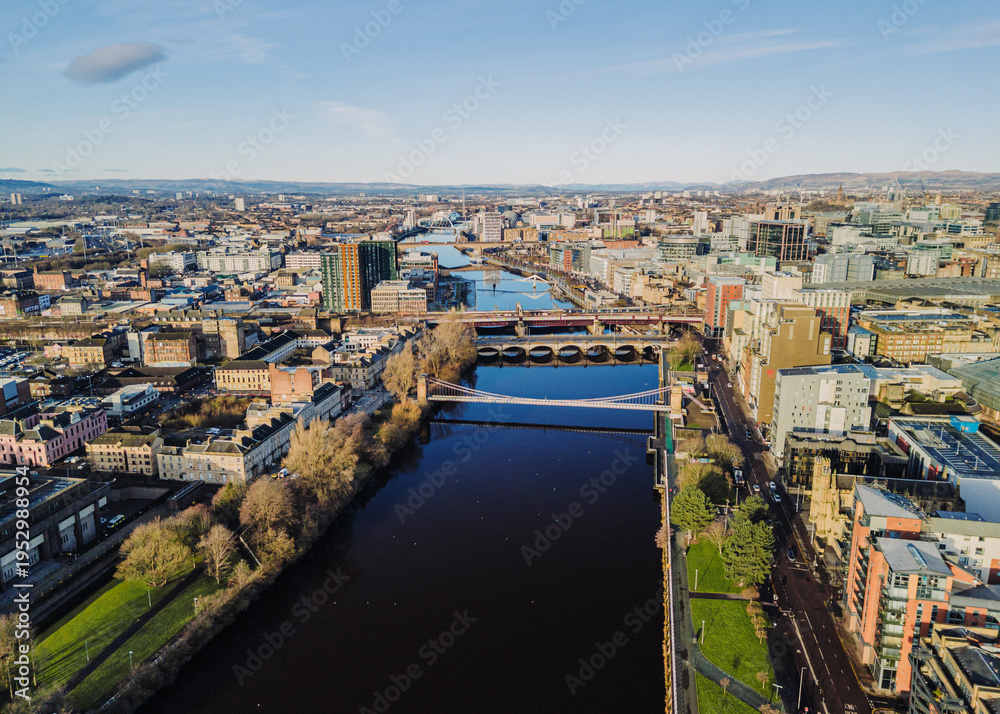 Obraz premium Glasgow Scotland: 13th Feb 2026: Caledonian Railway Bridge, King George V Bridge, Glasgow Bridge over River Clyde, with buildings lining the waterfront. Drone view golden hour with city skyline