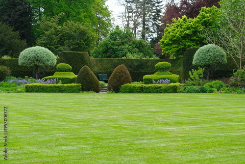 Manicured green lawn leads to a formal garden with ornate topiary and a secluded park bench against a dense hedge, embodying luxury landscaping, horticultural beauty, and serene estate elegance.