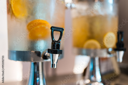 Refreshing beverages served from glass dispensers with taps for parties events. Large jars hold iced drinks with fruit slices like oranges, lemons. White cups stacked nearby for guests to use at