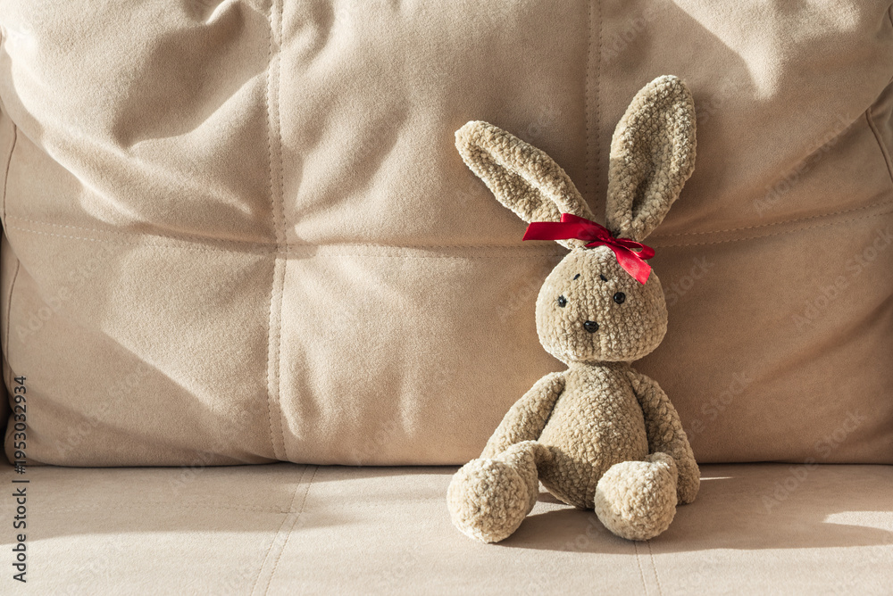 Fototapeta premium Plush Crocheted Bunny with Red Ribbon Sitting on Beige Couch