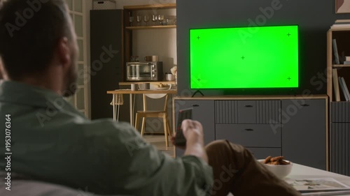 Medium rear shot of young man sitting on sofa in living room, switching channels with remote control while watching movie, show or sports match on TV with green screen with girlfriend or family