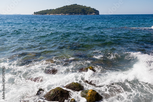 Scenic view of Lokrum Island from the rocky Adriatic coast in Dubrovnik, Croatia. Turquoise sea waves hitting mossy rocks under a clear summer sky. Beautiful Mediterranean coastal landscape.