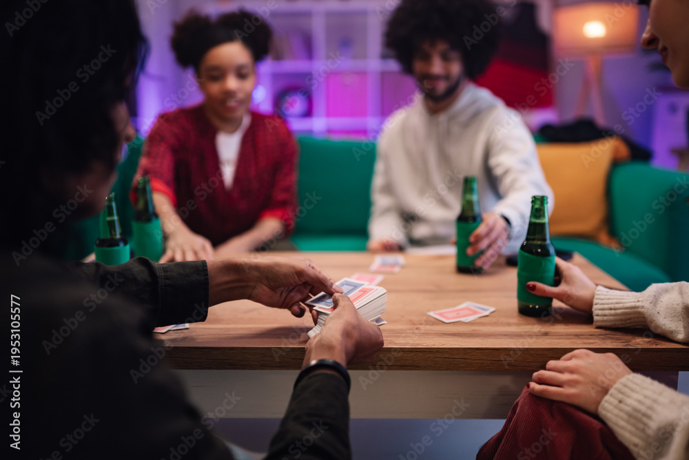 Fototapeta premium Diverse young adults friends playing cards and drinking beer in cozy living room enjoying social fun time together
