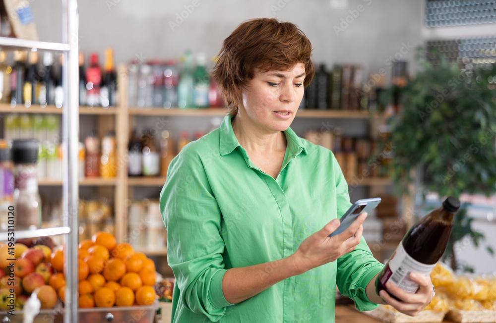 Fototapeta premium European woman scans the label on a bottle of pomegranate juice with her phone. Customer pays for purchases using a qr code and an application on phone