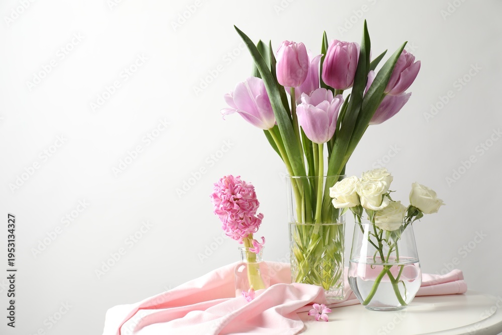 Fototapeta premium Beautiful flowers in vases on white table against light background. Space for text