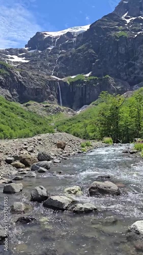 Vertical static shot of a clear mountain river flowing with a distant waterfall and glacier.