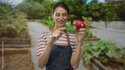 Woman in apron and gloves holding red apple, finger to lips for silence in forest garden beds; playful healthy eating.