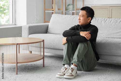 Canvastavla Sad young Asian man sitting on carpet at home