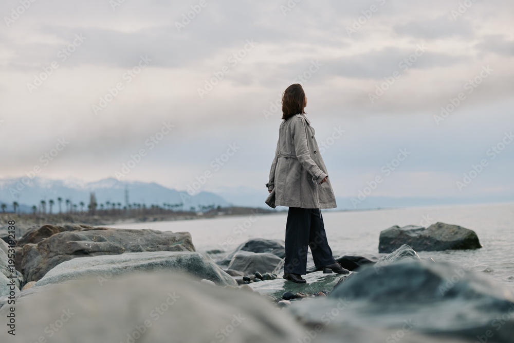 Fototapeta premium A woman in a checkered coat stands on rocks by a cloudy sea shore, looking into the distance. The scene evokes solitude, nature, and contemplation with muted colors and calm atmosphere.